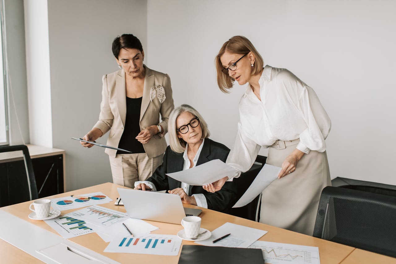 ROSARIUM CONSULTING Three businesswomen discussing reports in a modern office environment.
